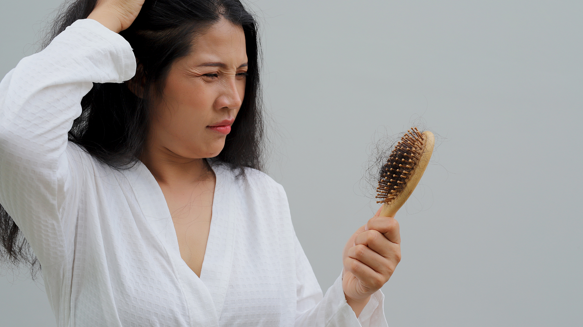 A woman holding a hair-filled brush while touching her hair, illustrating the reality of postpartum hair loss new moms face