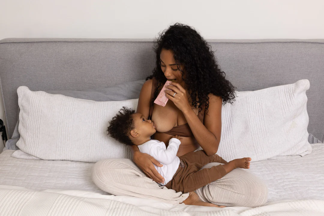 Mom breastfeeding her toddler on the bed while enjoying a BOOBIE lactation bar for on-the-go milk supply support.