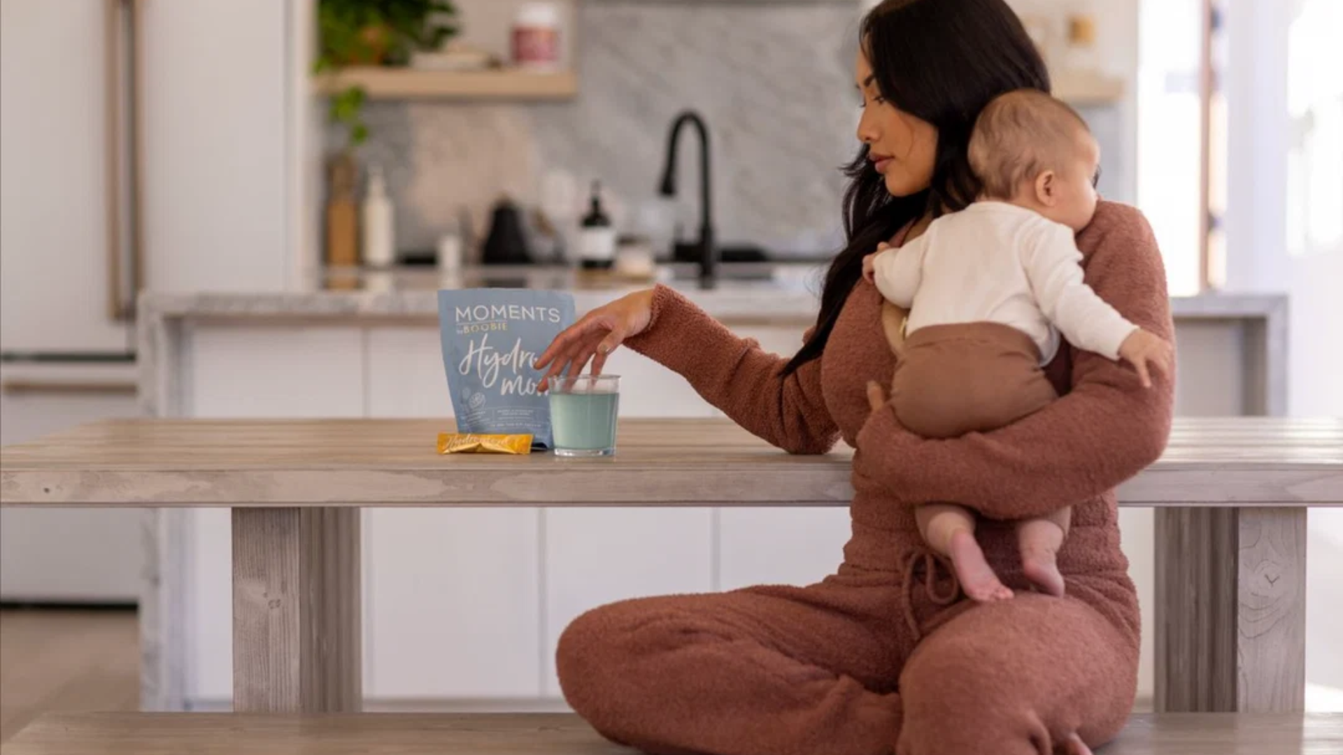 Mom holding her baby while reaching for a BOOBIE Hydromom drink.