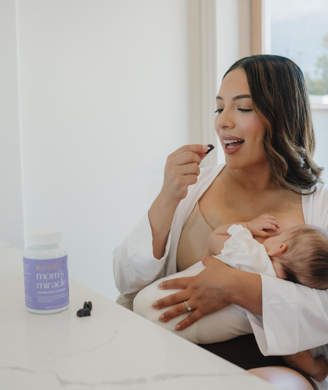 Woman breastfeeding a baby while holding a bottle labeled 'Mom's Miracle' in a bright room.