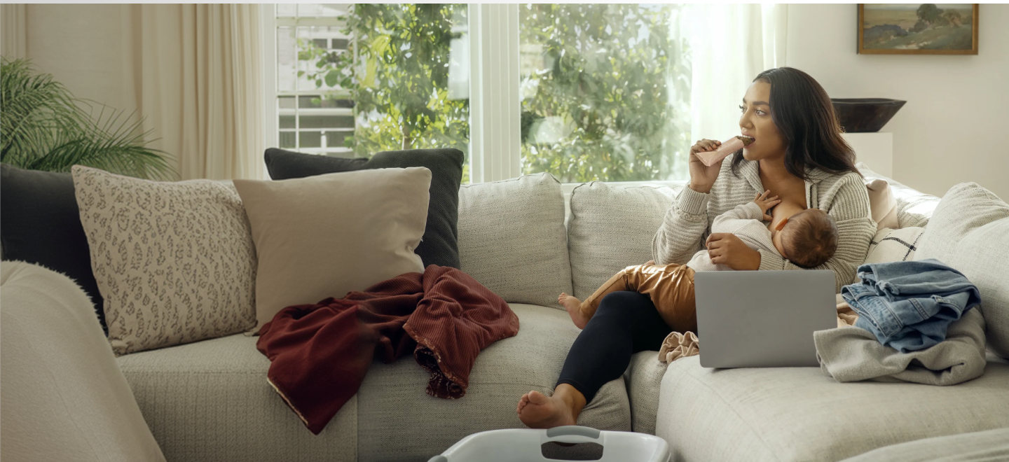 Woman holding a baby and breastfeeding while eating a Milk to the Max Lactation Bar from Boobie* Superfoods. Working on her laptop and folding clothes
