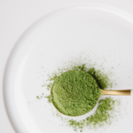 A close-up of bright green greens powder piled in a small gold spoon on a white plate, with a few loose green specks scattered nearby.
