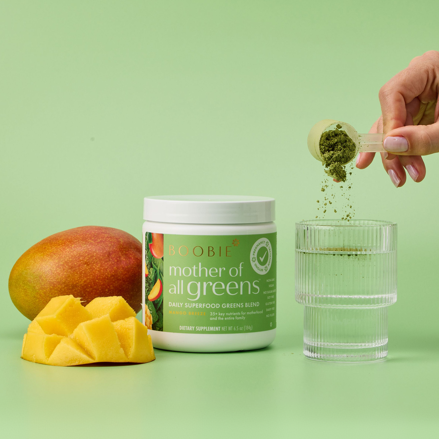 A hand holds a small scoop of green powder (Mother of All Greens Daily Superfood Greens Blend) above a clear ribbed glass, with the powder falling into the cup. The main product, a white tub labeled 'BOOBIE mother of all greens' and 'Daily Superfood Greens Blend,' stands beside a mango and sliced mango on a light green background.